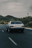 A silver Mercedes-Benz car is driving on an empty road with clear lane markings. The landscape features hills and sparse vegetation typical of a rural or semi-desert area. The sky is overcast, suggesting an approaching or passing storm.