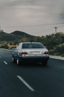 A silver Mercedes-Benz car is driving on an empty road with clear lane markings. The landscape features hills and sparse vegetation typical of a rural or semi-desert area. The sky is overcast, suggesting an approaching or passing storm.