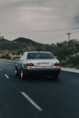 A silver Mercedes-Benz car is driving on an empty road with clear lane markings. The landscape features hills and sparse vegetation typical of a rural or semi-desert area. The sky is overcast, suggesting an approaching or passing storm.