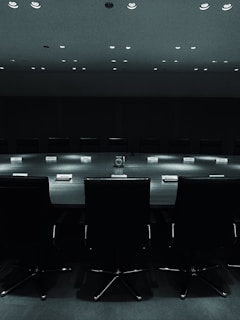 A large, dark conference room with a long table and empty chairs arranged around it. The lights above cast a subtle glow on the table, which has nameplates indicating seating arrangements. The mood is formal and slightly intense, emphasizing a lack of human presence.