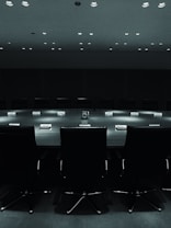 A large, dark conference room with a long table and empty chairs arranged around it. The lights above cast a subtle glow on the table, which has nameplates indicating seating arrangements. The mood is formal and slightly intense, emphasizing a lack of human presence.