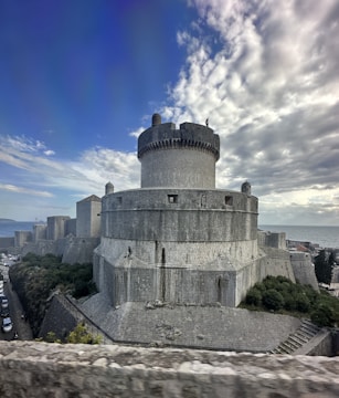 A large, circular stone fortress with thick walls and towers dominates the foreground. The structure exhibits medieval architectural features and is bathed in soft, overcast lighting from the cloudy sky above. Lush greenery surrounds some areas of the fortress, and a coastline is visible in the background.