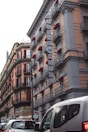 A street view featuring a large building with ornate architectural details and several balconies, each with red awnings. The signage for 'Airone Hotel' is prominently displayed on the building. In the foreground, there are several cars on the street.