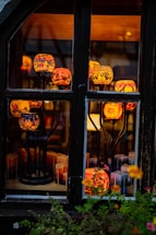 A collection of colorful, intricately designed candle holders displayed behind a window. The candle holders are elevated on black metal stands and emit a warm, inviting glow. Various patterns are visible on the holders, including urban landscapes and abstract designs. In the foreground, there are vibrant flowers, enhancing the cozy and artistic atmosphere.