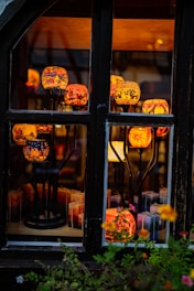 A collection of colorful, intricately designed candle holders displayed behind a window. The candle holders are elevated on black metal stands and emit a warm, inviting glow. Various patterns are visible on the holders, including urban landscapes and abstract designs. In the foreground, there are vibrant flowers, enhancing the cozy and artistic atmosphere.