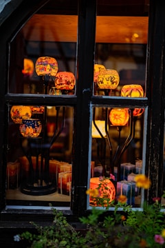 A collection of colorful, intricately designed candle holders displayed behind a window. The candle holders are elevated on black metal stands and emit a warm, inviting glow. Various patterns are visible on the holders, including urban landscapes and abstract designs. In the foreground, there are vibrant flowers, enhancing the cozy and artistic atmosphere.