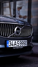 A close-up view of the front grille of a Volvo car, prominently displaying the brand's emblem. The car has a Turkish license plate, and a reflection of a person wearing a hard hat is visible in the background.