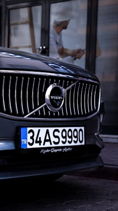 A close-up view of the front grille of a Volvo car, prominently displaying the brand's emblem. The car has a Turkish license plate, and a reflection of a person wearing a hard hat is visible in the background.