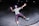A person wearing protective gear is skateboarding in a concrete skate park at night. The individual is mid-action, with arms outstretched and an expression of excitement or joy on their face. They are wearing a purple helmet, knee pads, elbow pads, and green sneakers.