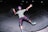 A person wearing protective gear is skateboarding in a concrete skate park at night. The individual is mid-action, with arms outstretched and an expression of excitement or joy on their face. They are wearing a purple helmet, knee pads, elbow pads, and green sneakers.