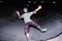 A person wearing protective gear is skateboarding in a concrete skate park at night. The individual is mid-action, with arms outstretched and an expression of excitement or joy on their face. They are wearing a purple helmet, knee pads, elbow pads, and green sneakers.