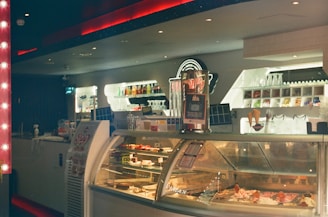 A cozy indoor gelato shop with a well-lit counter displaying various flavors of gelato in a refrigerated case. The background features shelves stocked with toppings and glassware. A sign with information is visible atop the counter.