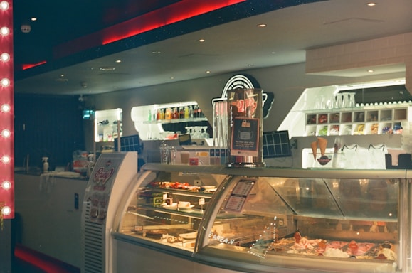 A cozy indoor gelato shop with a well-lit counter displaying various flavors of gelato in a refrigerated case. The background features shelves stocked with toppings and glassware. A sign with information is visible atop the counter.