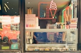 A display window of an ice cream and smoothie shop with various signs and price lists. The window showcases ice cream cones, neon lights in the shape of an ice cream cone, and several bottles of drinks. The words 'Gelato' and 'Smoothies Sold Here' are prominently displayed. The reflection of a red double-decker bus is visible in the glass.
