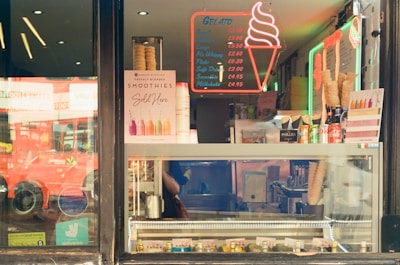 A display window of an ice cream and smoothie shop with various signs and price lists. The window showcases ice cream cones, neon lights in the shape of an ice cream cone, and several bottles of drinks. The words 'Gelato' and 'Smoothies Sold Here' are prominently displayed. The reflection of a red double-decker bus is visible in the glass.