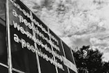 Metallic letters are affixed to a mesh surface creating a message that emphasizes the importance of positive vision and action. The scene is set against a backdrop of a partly cloudy sky, with boughs of trees visible at the bottom right.