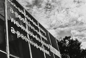 Metallic letters are affixed to a mesh surface creating a message that emphasizes the importance of positive vision and action. The scene is set against a backdrop of a partly cloudy sky, with boughs of trees visible at the bottom right.