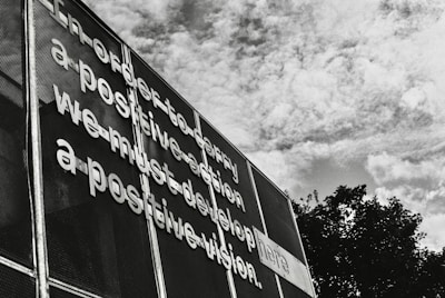 Metallic letters are affixed to a mesh surface creating a message that emphasizes the importance of positive vision and action. The scene is set against a backdrop of a partly cloudy sky, with boughs of trees visible at the bottom right.