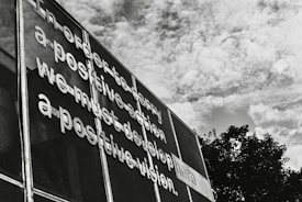 Metallic letters are affixed to a mesh surface creating a message that emphasizes the importance of positive vision and action. The scene is set against a backdrop of a partly cloudy sky, with boughs of trees visible at the bottom right.
