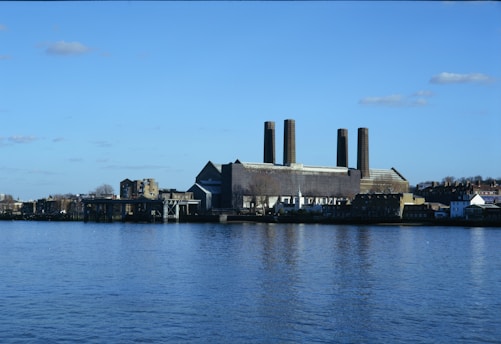 A large industrial building with four tall chimneys situated on the bank of a wide river. The structure resembles a power plant or factory. In the background, there are smaller buildings and a clear blue sky with a few scattered clouds.