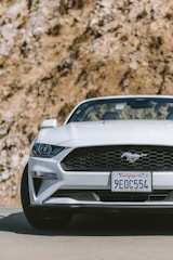 A white sports car with a distinctive emblem on the front grille is parked on a road. The license plate indicates it's from California. The background features a rocky surface with some visible vegetation.