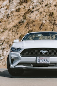 A white sports car with a distinctive emblem on the front grille is parked on a road. The license plate indicates it's from California. The background features a rocky surface with some visible vegetation.