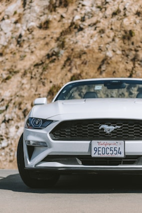 A white sports car with a distinctive emblem on the front grille is parked on a road. The license plate indicates it's from California. The background features a rocky surface with some visible vegetation.