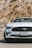 A white sports car with a distinctive emblem on the front grille is parked on a road. The license plate indicates it's from California. The background features a rocky surface with some visible vegetation.
