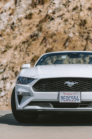 A white sports car with a distinctive emblem on the front grille is parked on a road. The license plate indicates it's from California. The background features a rocky surface with some visible vegetation.