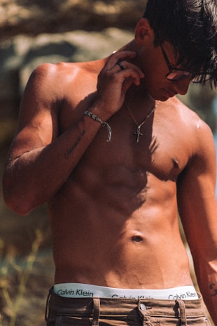 A shirtless man with a muscular build is wearing glasses and a necklace with a cross pendant. His hand rests near his face, and he has tattoos on his arm. He is wearing Calvin Klein underwear and brown pants.