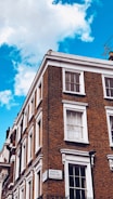 A corner of a brick building with multiple white-framed windows. The building features a classic architectural style with detailed cornices. A street sign on the building indicates 'Craven Road W2, City of Westminster.' The sky is a vibrant blue with a few white clouds.