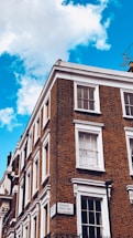A corner of a brick building with multiple white-framed windows. The building features a classic architectural style with detailed cornices. A street sign on the building indicates 'Craven Road W2, City of Westminster.' The sky is a vibrant blue with a few white clouds.