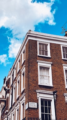 A corner of a brick building with multiple white-framed windows. The building features a classic architectural style with detailed cornices. A street sign on the building indicates 'Craven Road W2, City of Westminster.' The sky is a vibrant blue with a few white clouds.
