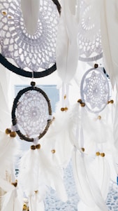 An intricate pattern of dreamcatchers hanging, featuring lacy designs and adorned with white feathers and small wooden beads. The dreamcatchers have circular frames wrapped in dark material, creating a contrast against the delicate white details.