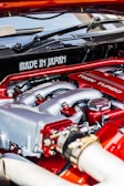 A close-up view of a car engine featuring prominently the words 'Made in Japan' and 'Twin Turbo'. The engine components are detailed, with visible pipes, bolts, and metal surfaces. The colors red and silver are dominant in the design, indicating performance and craftsmanship.