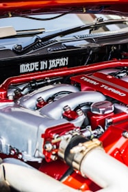 A close-up view of a car engine featuring prominently the words 'Made in Japan' and 'Twin Turbo'. The engine components are detailed, with visible pipes, bolts, and metal surfaces. The colors red and silver are dominant in the design, indicating performance and craftsmanship.