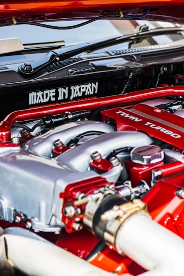 A close-up view of a car engine featuring prominently the words 'Made in Japan' and 'Twin Turbo'. The engine components are detailed, with visible pipes, bolts, and metal surfaces. The colors red and silver are dominant in the design, indicating performance and craftsmanship.