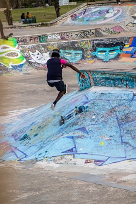 A skateboarder is captured mid-jump performing a trick over a graffiti-covered skate park obstacle. Vivid artwork and graffiti in greens, blues, and purples decorate the concrete structures, adding an urban and creative vibe. Nearby, a few people are sitting in the grassy area in the background.