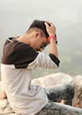 A person is sitting on a rocky surface, wearing a long-sleeved shirt and jeans, with one hand on their head. They are looking downward, and appear to be outdoors with a blurred natural backdrop.