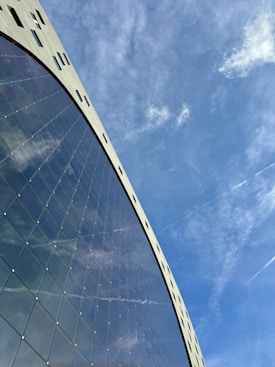 A modern architectural structure with a curved glass facade reflecting the sky. The building is designed with geometric precision, featuring a grid pattern on the glass and small windows at the top. Wispy clouds and contrails are visible in the bright blue sky.