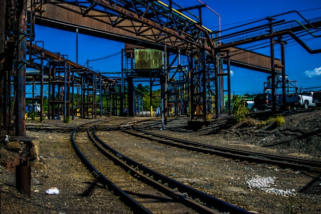 An industrial scene with a complex network of metal structures and railroad tracks curving through the area. There are steel beams and ladders forming part of the architecture, with a clear blue sky in the background. Several vehicles are parked along the side.
