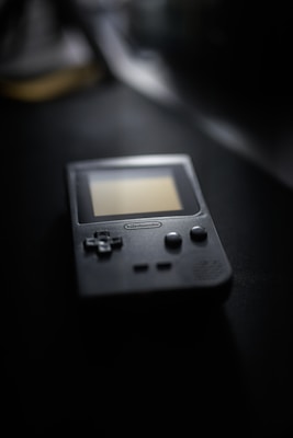 A vintage handheld gaming console with a screen and control buttons is placed on a dark surface. The focus is on the console, with the background blurred, creating a nostalgic atmosphere.