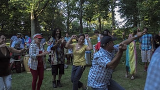 A diverse group of people are dancing and playing music in a park, surrounded by lush green trees. Some individuals are clapping, playing drums, or dancing energetically. The scene captures a lively and joyful atmosphere.