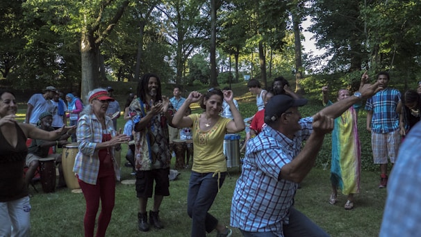 A diverse group of people are dancing and playing music in a park, surrounded by lush green trees. Some individuals are clapping, playing drums, or dancing energetically. The scene captures a lively and joyful atmosphere.