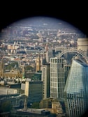 A cityscape featuring a blend of historical and modern architecture. Prominent landmarks include a well-known clock tower and a giant ferris wheel. The scene is observed from a window or through a lens, creating a circular vignette effect around the edges.