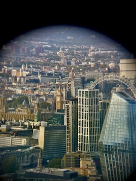 A cityscape featuring a blend of historical and modern architecture. Prominent landmarks include a well-known clock tower and a giant ferris wheel. The scene is observed from a window or through a lens, creating a circular vignette effect around the edges.