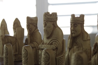 A group of intricately carved chess pieces made from stone or a stone-like material, featuring detailed human figures, some wearing crowns or hats. The pieces exhibit historical or medieval styling, suggesting they might be replicas of an ancient chess set. The lighting is soft, casting shadows that enhance the textures and expressions on the figures.