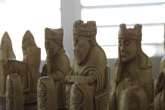 A group of intricately carved chess pieces made from stone or a stone-like material, featuring detailed human figures, some wearing crowns or hats. The pieces exhibit historical or medieval styling, suggesting they might be replicas of an ancient chess set. The lighting is soft, casting shadows that enhance the textures and expressions on the figures.