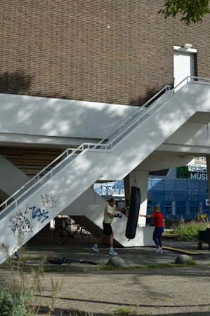 Two people are engaged in a boxing workout outdoors under a stairway. One person is punching a suspended punching bag while the other seems to be instructing or observing. The area has a mix of natural and urban elements, with greenery and a brick wall in the background, as well as bicycles and graffiti on the structure.