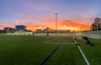 A soccer field is depicted during sunset, with an expansive sky featuring vibrant orange, pink, and purple hues. Players are scattered across the field, some practicing and others standing near the goalposts. The field is enclosed by a white fence, and in the background, modern buildings and trees are visible, illuminated by artificial lights.